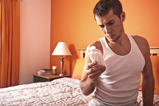 Man sits on his bed and looks pensively at a bottle of pills. Horizontal format.