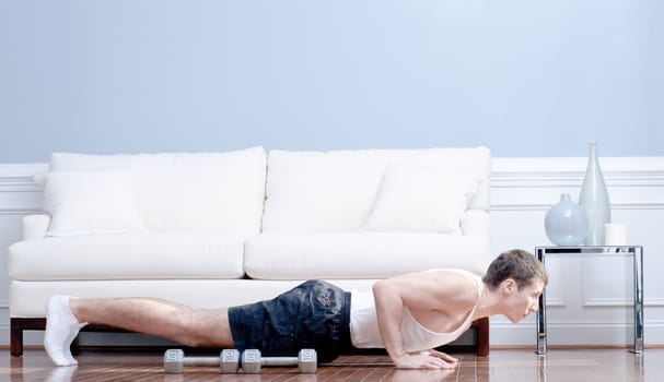 Full length view of man doing push-ups next to couch, with arm weights lying next to him. Horizontal format.