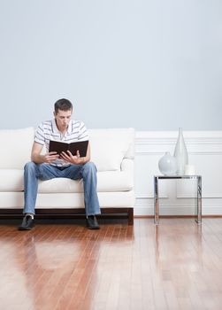 Man sitting and reading on a white couch in his living room. Vertical format.