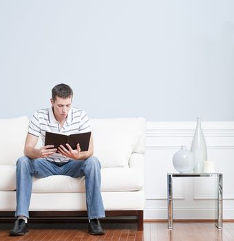 Man sitting and reading on a white couch in his living room. Square format.