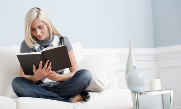 Woman sitting cross-legged on white couch and reading a book. Horizontal format.