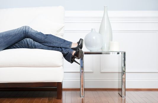 Cropped view of woman reclining on white couch next to an end table holding vases, with only her legs visible. Horizontal format.
