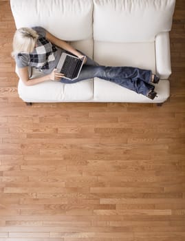 Full length overhead view of woman reclining on white couch and using a laptop. Vertical format.