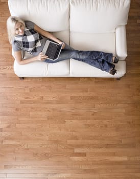 Full length overhead view of woman smiling up at the camera as she reclines on a white couch with a laptop. Vertical format.