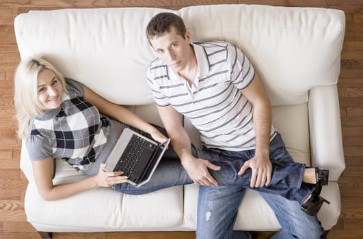 Cropped overhead view of couple relaxing together on white couch and looking up at camera, with woman using laptop and stretching out with her legs in the man's lap. Horizontal format.