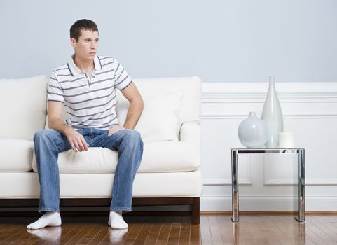 Man in casual clothing sitting on a white couch in a living room. He has a blank expression and is looking to the side. Horizontal format.