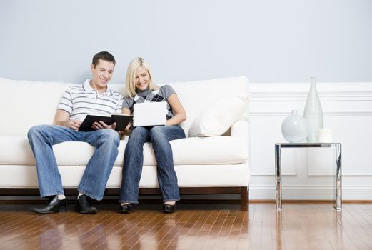 Man is reading, and woman is using a laptop, as they sit side by side on a white couch. Horizontal format.