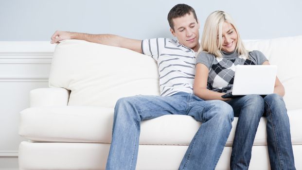 Man and woman sit side by side on a white couch. The woman is using a laptop, and the man is snuggling next to her. Horizontal format.