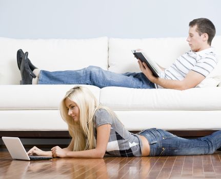 Man reads on a couch while woman stretches out on the floor with her laptop. Horizontal format.