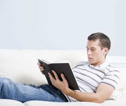 Cropped view of a man reclining on a white couch with a book. Horizontal format.