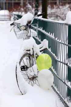 a bike covered with a lot of snow, standing at a fence, decorated with balloons