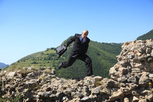 Businessman with a laptop bag running outdoors on a rocky ruined stronghold wall in a mountaineous area.The image can suggest the idea of a thief stealing important data or a man protecting data in case of a disaster.