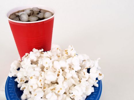 A blue bowl of fluffy white popcorn and a red cup with soda in it, over a white background