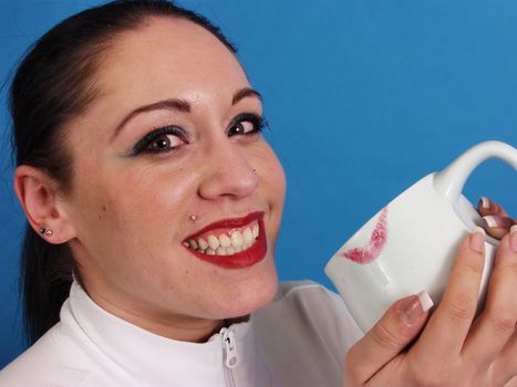 A beautiful brunette smiles broadly for the camera, a coffee cup with a deep red lip print on the rim is cradled in her hands. On a blue background.