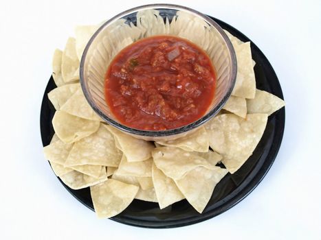 A bowl of salsa on a black plate full of chips. Over a white background.