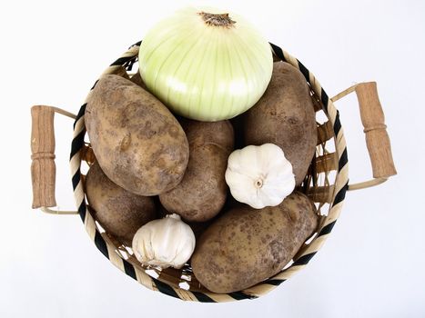 A wicker basket full of baker potatoes, a large white onion, and garlic bulbs. Over a white background.