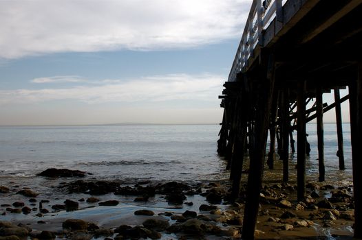 From ground level along the side of a rickety wooden pier, POV, into the ocean with rocks and sand low down, the pier struts and a cloudy sky in the distance