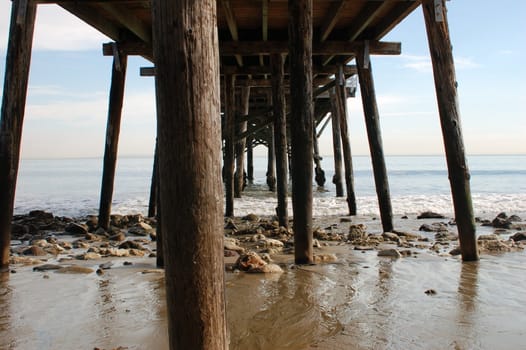 View out to sea from the underside of an old wooden pier/ jetty with full view of many wooden struts into the sand, rocks and the sea and sky merging in the distance