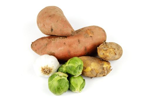 Two raw unpeeled sweet potatoes with a bulb of garlic, green sprouts and potatoes on a reflective white background