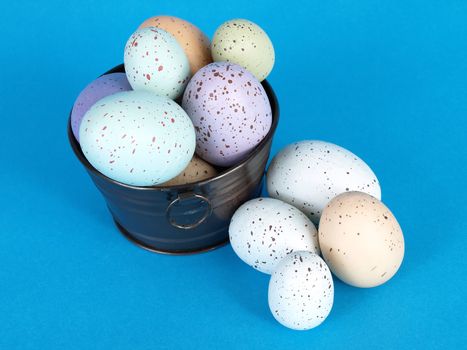 Flecked pastel colored eggs fill a metal pail, a few eggs laying to the side, over a blue background.