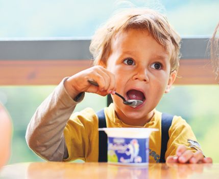 Little boy is eating yogurt, holding a spoon in his hand, sitting at the table .