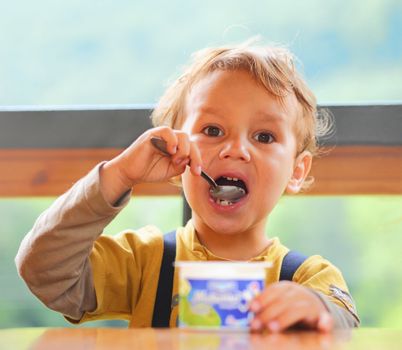 Little boy is eating yogurt, holding a spoon in his hand, sitting at the table .