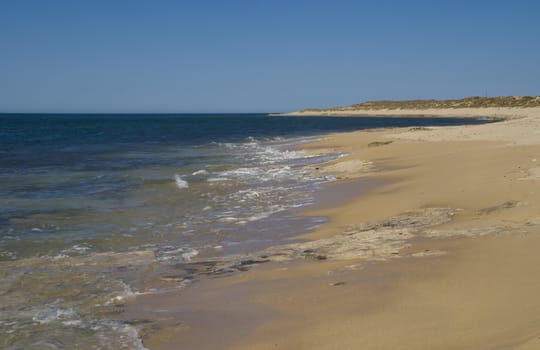 A deserted beach, composed of flat rocks and sand, as a deserted setting in the evening. Dunes are set in a distance against the blue sky. 