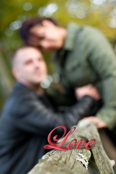 A young happy couple outdoors out of focus with the word LOVE in the foreground resting on the fence post.  Shallow depth of field.