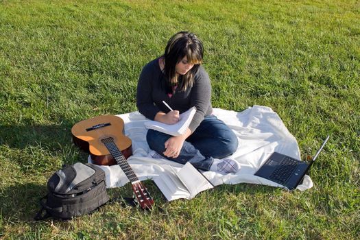 A young female singer or song writer with her guitar and computer outdoors in the grass.