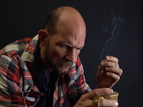 An older man sits, drinking coffee and smoking a cigarette, a contemplative or worried look on his face.