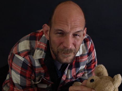 An older balding male holds a teddy bear, tears running down his face.