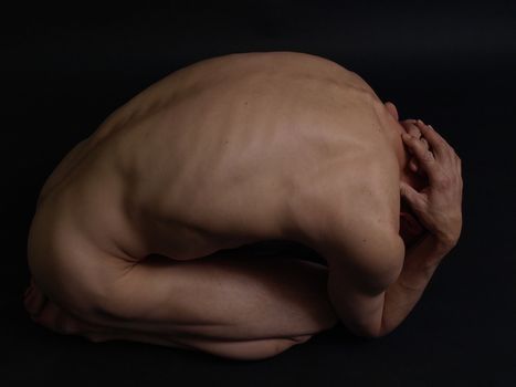 A nude male curled up into a ball on a black background.