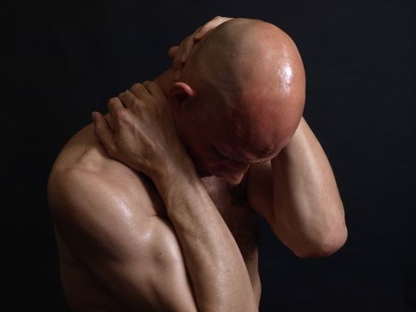 A shirtless bald male adult grips at his head and neck over a black background.