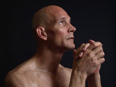 A bald shirtless man clasps his hands in prayer, eyes pleading skyward, over a black background.