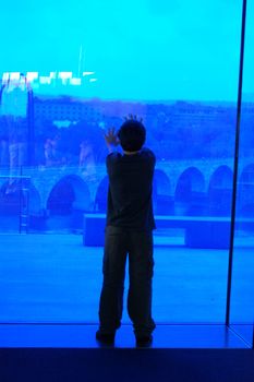 Boy looking through blue glass window onto city below.
