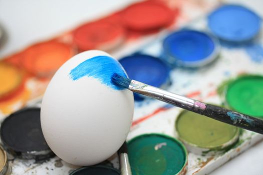 A white egg with some blue paint and a pencil, other colors of paint in the background