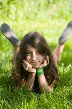 The smiling dark-haired girl-teenager lays on a green lawn in sunlight patches
