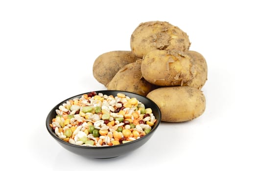 Small pile of brown unpeeled potatoes with a small black dish of soup pulses on a reflective white background