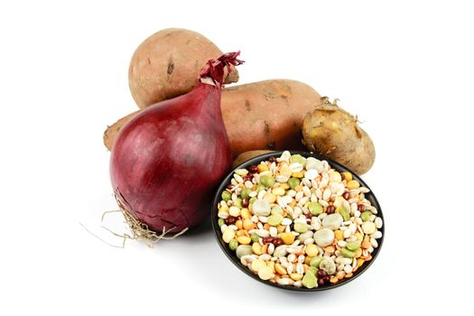 Two raw unpeeled sweet potatoes with a red onion and a black dish of soup pulses on a reflective white background