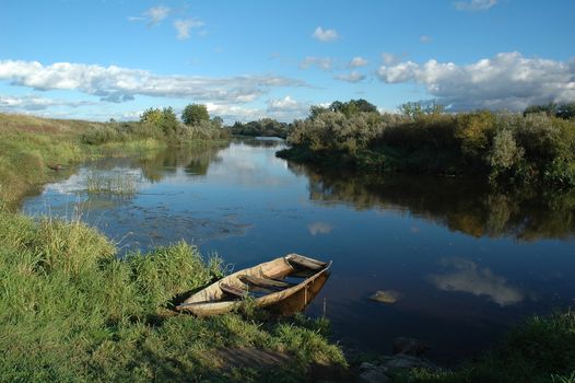 Tranquil, relaxing panorama from the bank of a quiet Russian river with a row boat partially submerged in the foreground and beautiful clouds in a blue sky.