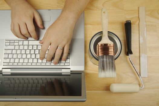 Handyman Researches on Laptop with paint brush, roller and wood stir stick by his side. Great image for online information regarding home improvement, additions and remodeling.