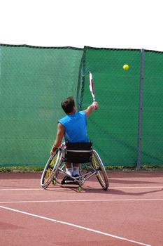 A wheelchair tennis player during a tennis championship match, taking a shot. Space for text above the fence.