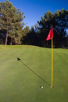 Golf hole with red flag, golf ball and shadow.