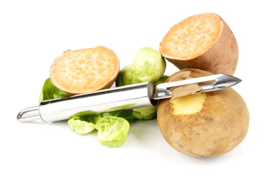 Small brown raw potato with chrome peeler and a sweet potato cut in half with sprouts on a reflective white background