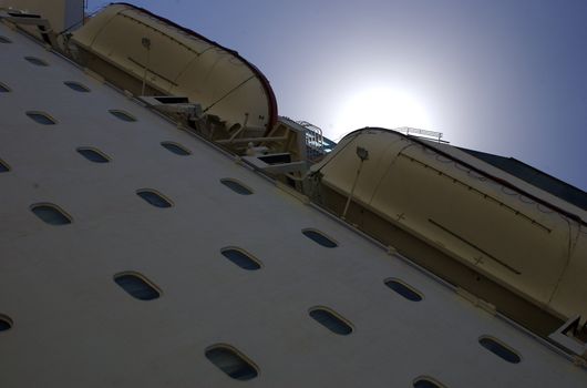 View of a tall cruise liner taken from below up the side into the sky, with a view of the sun peeking out above the ship in the centre of the shot