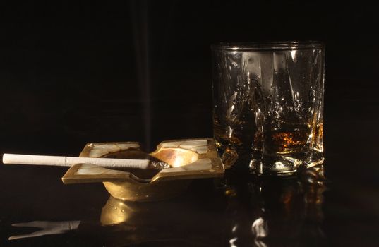 Old bronze ash-tray with cigarette and glass of whisky on dark background