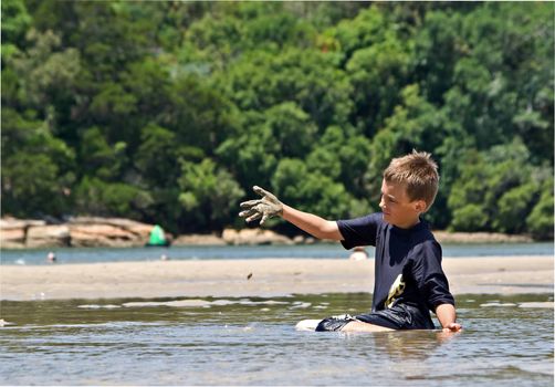 a boy sitting the water lifts a handful of mud