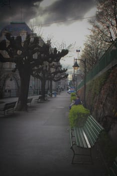 Image of a sidewalk with park benches and an unrecognizable woman reading a newspaper under a street lamp.
