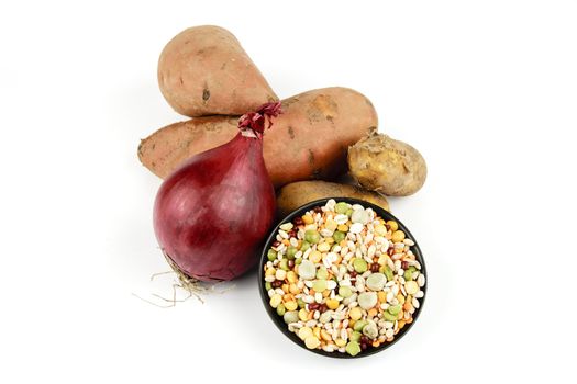 Two raw unpeeled sweet potatoes with a red onion and a black dish of soup pulses on a reflective white background