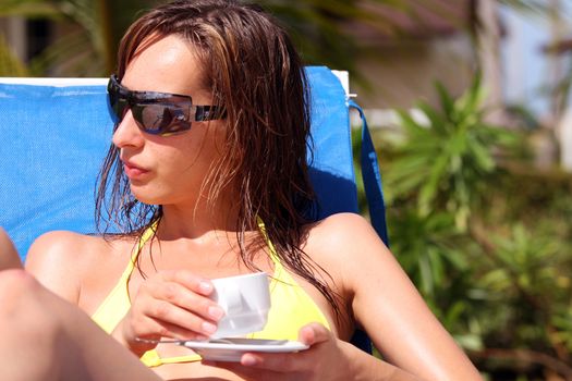 woman sitting in chaise and drinking cup coffee by the pool